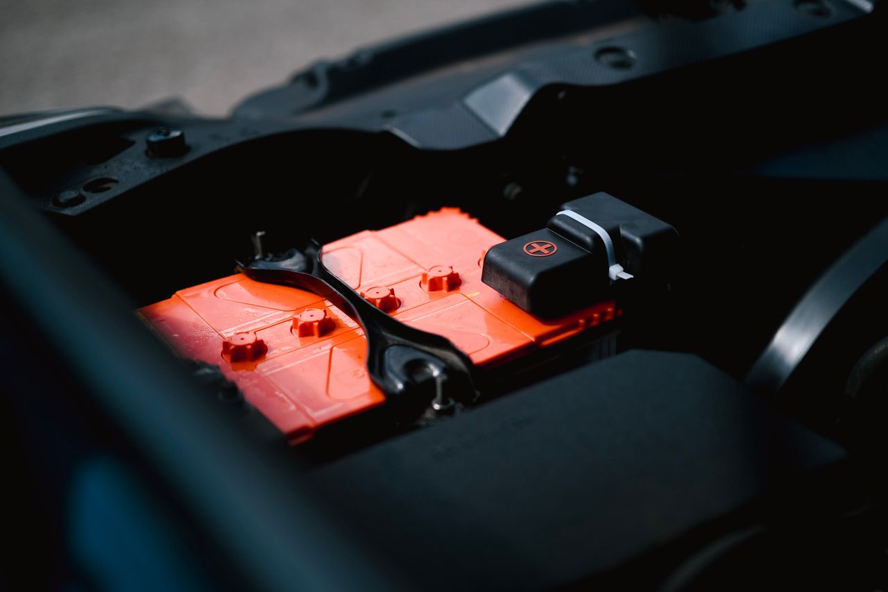 our-story-01 Detailed view of an orange car battery inside a vehicle's engine bay, highlighting its features.